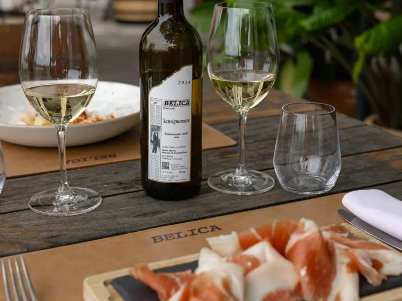 {"sl":"Steklenica vina Belica in dva kozarca belega vina na leseni mizi, v ozadju kro\u017enik in narezek pr\u0161uta v ospredju.","en":"","de":"","it":""}